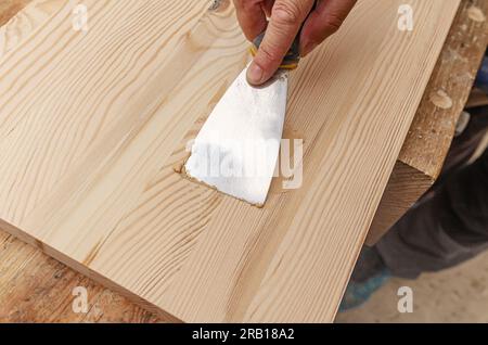 Couvrir les nœuds sur une planche de bois avec du mastic. Spatule, mastic, planche de bois. Les mains de Carpenter. Banque D'Images