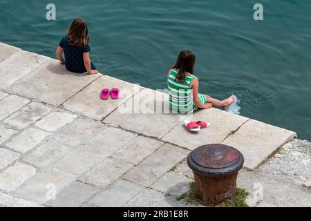 deux jeunes enfants assis sur un mur de mer plongeant les pieds dans l'eau, des jeunes filles pagayant au bord de la mer, deux jeunes filles rafraîchissant leurs pieds dans l'eau. Banque D'Images