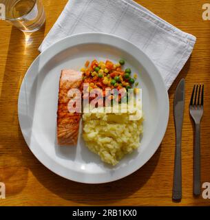 Saumon grillé avec pommes de terre écrasées et légumes sur une assiette blanche couchée sur une table en bois, nappe blanche et verre d'eau. Vue de dessus. Banque D'Images