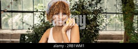 Heureuse jeune femme afro-américaine avec des bretelles portant le foulard et la robe d'été et tenant le citron frais et debout dans l'orangerie floue, élégant Banque D'Images