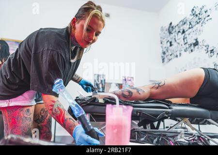 Jeune fille tatoueuse caucasienne dessinant un tatouage sur la jambe d'un client, salon de tatouage plein plan moyen. Photo de haute qualité Banque D'Images