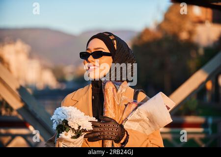 Une hijab femme dans des lunettes de soleil élégantes et une élégante tenue française, marchant dans la ville au coucher du soleil, portant un bouquet, du pain, et un journal Banque D'Images