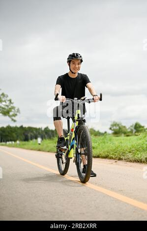 Un portrait d'un beau jeune homme asiatique en vêtements de sport monte un vélo sur les routes de campagne. Un cycliste masculin qui roule le long de la rue. Banque D'Images