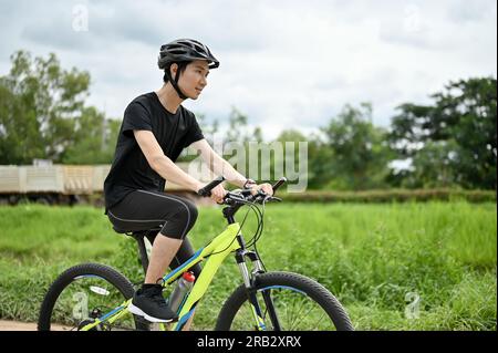 Un homme asiatique heureux et en bonne santé en vêtements de sport et un casque de vélo monte un vélo le long des routes de campagne. concept d'activité estivale Banque D'Images