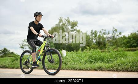 Un homme asiatique déterminé et en bonne santé en vêtements de sport et un casque de vélo monte un vélo le long des routes de campagne. sport, exercice, loisirs, activité estivale Banque D'Images