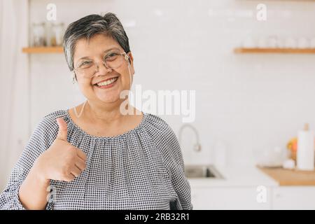 portrait heureuse femme asiatique mature en bonne santé souriante mignonne belle à la maison cuisine main pouces vers le haut Banque D'Images