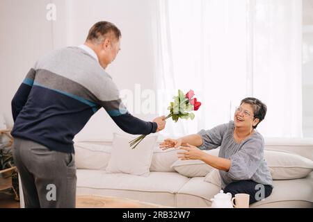 couple adulte senior amoureux mignon doux surprise donnant des fleurs de roses pour l'anniversaire de mariage ou les jours de saint-valentin à sa femme à la maison Banque D'Images