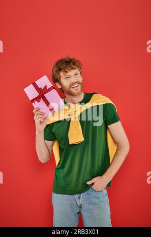 vacances, homme roux heureux avec barbe posant dans une tenue décontractée sur fond de corail, tenant boîte-cadeau, occasions festives, cadeau emballé, mode et t Banque D'Images