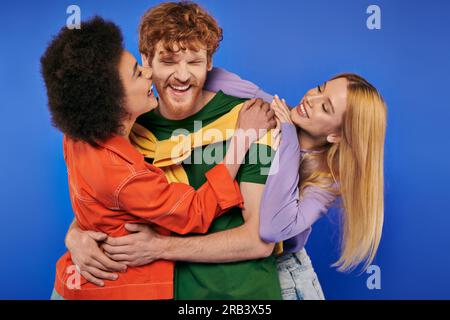 relations alternatives, amateurs de polyamory, femmes multiethniques gaies embrassant et embrassant un jeune homme rousse sur fond bleu, photo en studio, vibrant Banque D'Images