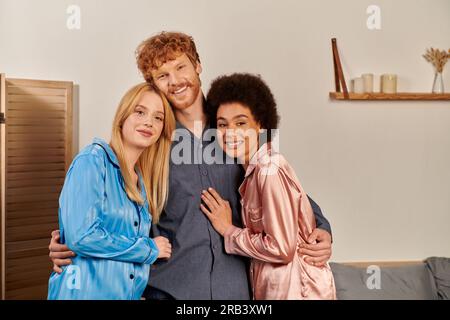 amateurs de polyamory en pyjama, homme heureux avec les cheveux roux embrassant les femmes interraciales à la maison, diversité culturelle, partenaires non traditionnels, liberté en relat Banque D'Images