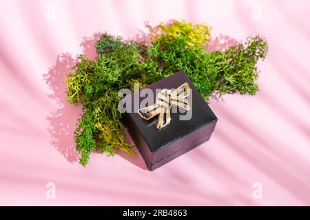 Broche dorée en forme d'arc sur une boîte à bijoux noire à côté de lichen vert sur fond rose avec des ombres de feuilles de palmier, gros plan Banque D'Images