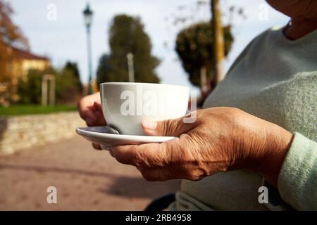 Les mains d'une femme âgée tenant une tasse de café à l'extérieur par une journée ensoleillée d'automne Banque D'Images