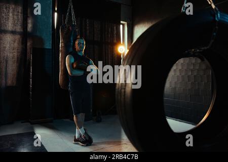 Boxer fatigué dans des gants de boxe reposant dans la salle de gym sombre après l'exercice près du sac de frappe et du pneu. Banque D'Images