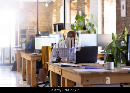Heureux homme d'affaires afro-américain occasionnel utilisant un ordinateur portable et déjeunant au bureau. Affaires, nourriture, technologie et travail, inchangés. Banque D'Images