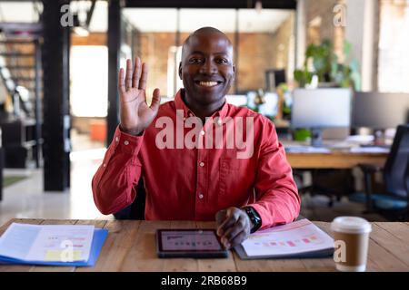 Portrait de heureux homme d'affaires afro-américain occasionnel ayant un appel vidéo et agitant la main dans le bureau. Affaires, technologie et travail, inchangés. Banque D'Images