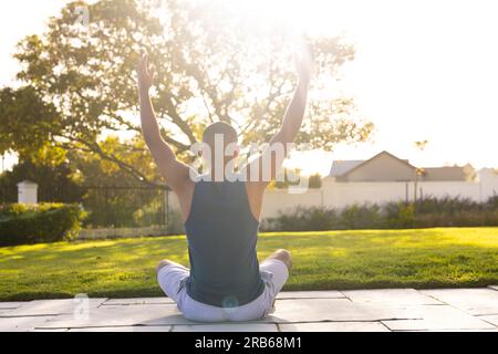 Vue arrière de l'homme biracial pratiquant le yoga assis dans le jardin ensoleillé. Été, bien-être, fitness et mode de vie sain, inchangés. Banque D'Images