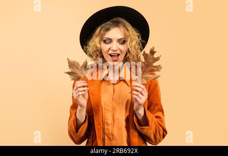 Fille surprise dans le chapeau noir avec des feuilles d'automne. Femme de mode en chemise orange et manteau avec feuillage automnal. Soldes d'automne. Remise. Vêtements d'automne Banque D'Images