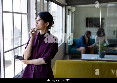 Femme d'affaires asiatique décontractée réfléchie debout regardant par la fenêtre dans le bureau, espace de copie. Travail, affaires, inspiration et bureau décontracté, inchangé. Banque D'Images