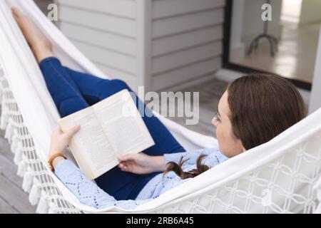 Femme caucasienne détendue couchée dans un hamac et livre de lecture dans le jardin. Vie domestique, relaxation et mode de vie, inchangés. Banque D'Images
