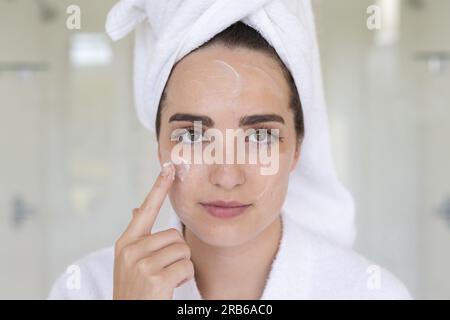 Portrait de femme caucasienne avec une serviette sur la tête appliquant de la crème sur le visage dans la salle de bain. Vie domestique, soins personnels et mode de vie, inchangés. Banque D'Images