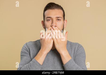 Je ne dirai personne. Jeune homme libanais effrayé fermant la bouche avec la main, l'air intimidé effrayé à la caméra, des gestes non, refusant de dire terrible secret incroyable vérité sur fond beige Banque D'Images