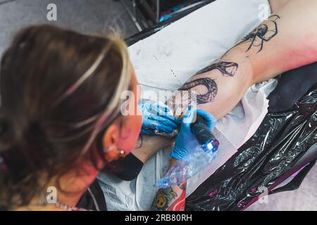 Artiste tatoueur POV. Tatoueuse féminine dessinant un tatouage de dragon ou de serpent sur un mollet de son client à l'aide d'aiguilles professionnelles et de gants de protection. Photo de haute qualité Banque D'Images