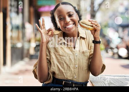 Burger, signe d'accord et portrait de femme dans la ville, restaurant en plein air et expérience client heureux ou services. La restauration rapide, comme et excitée africaine Banque D'Images