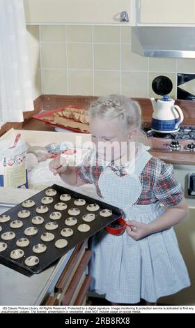 Au début des années 1960 Une fille joliment habillée dans un tablier met des raisins secs sur le dessus des biscuits presque prêts à cuire. Il semble que c'est proche de noël car il y a déjà un plateau de pain d'épices prêt à l'emploi sur le comptoir. L'intérieur de la cuisine est typique de la décennie. Suède 1960 Banque D'Images