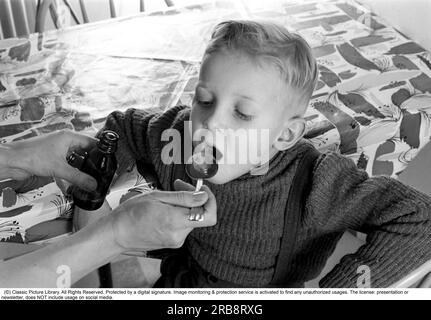 Un garçon est malade et refuse de prendre ses médicaments. Sa mère tient une cuillère et attend qu'il gape et la prenne, et il le fait. Il ouvre sa bouche et le prend même si elle ne tase pas si bien. Suède 1956. Conard réf. 3157. Banque D'Images