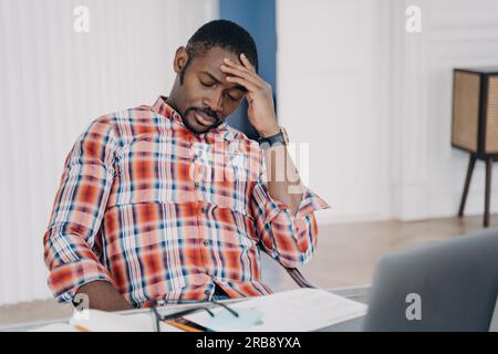 Homme d'affaires afro-américain fatigué souffrant de maux de tête, après avoir travaillé sur ordinateur portable, assis sur le lieu de travail. Homme noir surmené touchant la tête, po Banque D'Images