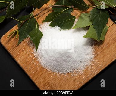 Édulcorant utile Xylitol, sucre de bouleau sur planche de bois à découper, bâtons, feuilles de bouleau, fond sombre. Nutriment sain, alimentation. Pose à plat. Horizontal Banque D'Images