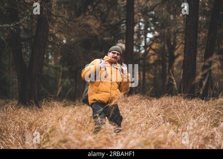 Jeune homme avec veste d'hiver jaune et chapeau, mains dans les poches, se tient dans de l'herbe haute jaune et sèche, pendant l'automne froid jour nuageux, Decin, la réputation tchèque Banque D'Images