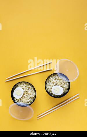 Deux bols en plastique de nouilles instantanées avec des œufs sont servis avec des baguettes plates sur fond de papier jaune avec espace de copie Banque D'Images