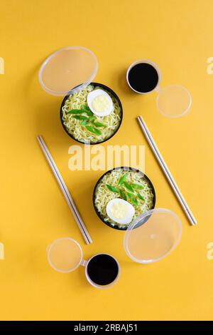 Deux bols en plastique de nouilles instantanées avec des œufs pondus à plat sont servis avec des baguettes et de la sauce soja sur fond de papier jaune. Banque D'Images