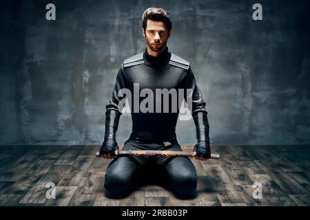 Homme guerrier assis sur le sol posant avec une épée dessus arrière-plan noir Banque D'Images