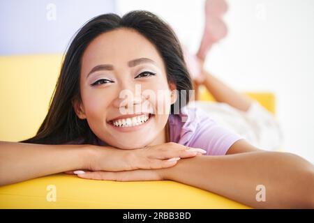 Gros plan heureuse femme asiatique portrait se relaxant à la maison allongé sur un canapé jaune. Repos, beauté féminine Banque D'Images