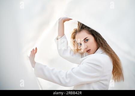 Jeune femme attrayante en vêtements blancs posant sur un arrière-plan abstrait pâle regardant l'appareil photo confiant.Couleurs douces et ensoleillées.Concept de beauté Banque D'Images