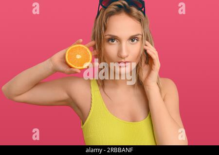 Jolie jeune femme blonde avec des lunettes de soleil sur la tête tenant une orange fraîche avec une coupe de moitié sur sa joue alors qu'elle regarde soigneusement la caméra sur un Banque D'Images