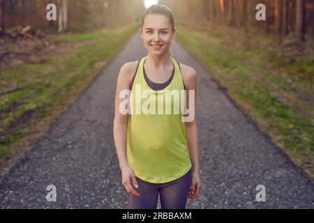 Jeune femme souriante en forme athlétique s'exerçant sur une voie goudronnée à travers les forêts rétro-éclairées par la lueur chaude du soleil dans un mode de vie actif sain Banque D'Images