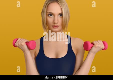 Jolie femme blonde en top d'entraînement noir avec de petits haltères roses dans ses mains, regardant la caméra. Gros plan portrait avant sur jaune ordinaire Banque D'Images