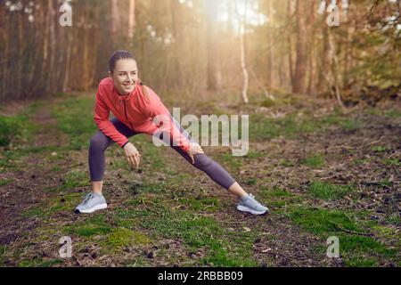 Portrait en longueur d'une jeune femme en forme heureuse faisant des exercices d'étirement pour les jambes et les bras pendant l'entraînement en plein air dans la forêt au printemps Banque D'Images