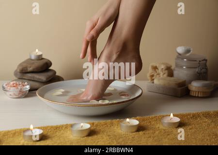 Femme trempant son pied dans un bol avec de l'eau et des pétales sur plancher en bois blanc, gros plan. Procédé de pédicure Banque D'Images