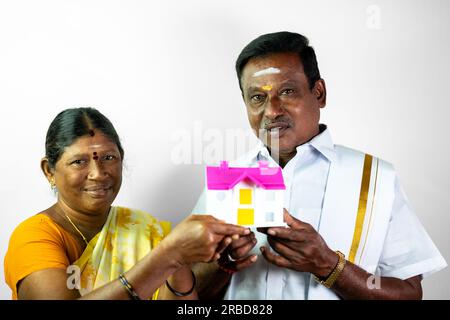 Un mari et une femme indiens se tiennent devant une caméra, tenant une petite maison de jouets. Le mari sourit et pointe le doigt vers la maison, la femme l'est Banque D'Images