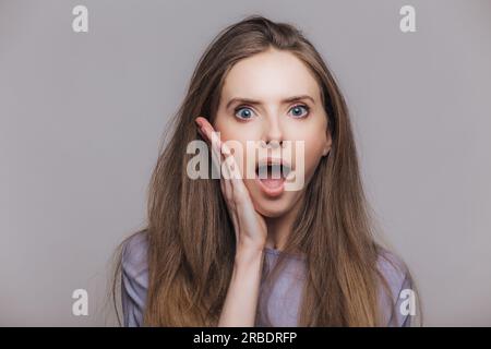 Femme brune émerveillée avec la bouche grande ouverte et l'expression stupéfaite. Surpris par des nouvelles inattendues. Fond de studio gris. Banque D'Images