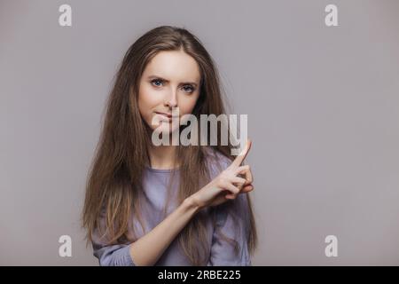 Mignon modèle brunette pointe sur le mur gris blanc, annonce avec une expression sérieuse. Concept publicitaire attirant l'attention. Banque D'Images