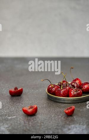 Quelques délicieuses cerises mûres dans une petite soucoupe en métal bordée d'or, certaines coupées sur une surface sombre Banque D'Images