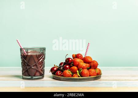 Un verre de jus de détox de baies rouges avec une paille, des baies rouges mûres sur une table de couleur claire et un fond bleu Uni Banque D'Images