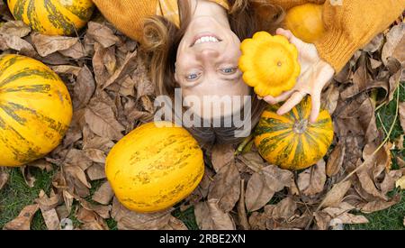 femme souriante heureuse se trouve sur un tas de feuilles sèches parmi les grandes et petites citrouilles, l'une d'elles tient en main près du visage. mise au point sélective, vue de dessus. Beaut Banque D'Images
