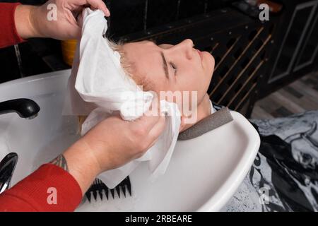 Les mains d'un barbier professionnel essuyant les cheveux sur la tête du client d'un jeune gars au-dessus du, coulent avant une coupe de cheveux dans le salon. Banque D'Images