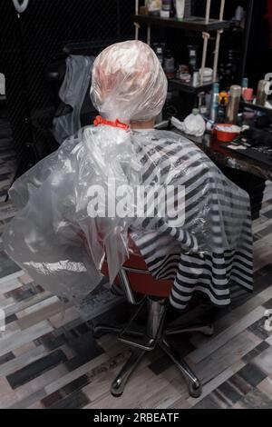 Une fille avec un film fixe sur sa tête est assise dans une chaise de salon de coiffure dans le processus de teinture de ses cheveux et de la tête avec des services de salon de peinture. Banque D'Images